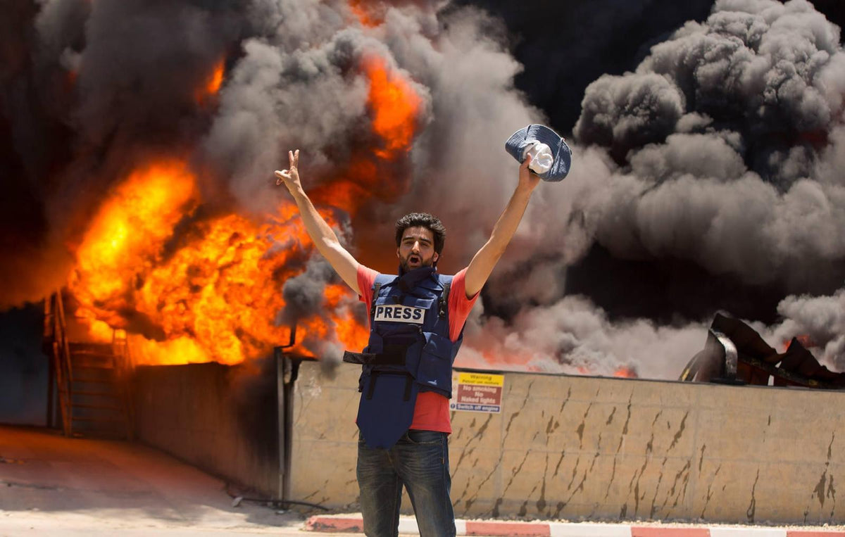 Wissam Nassar reporting in the field, wearing a press vest, in front of a large fire and smoke